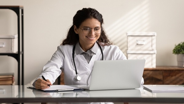 Happy young female doctor taking notes while using laptop.